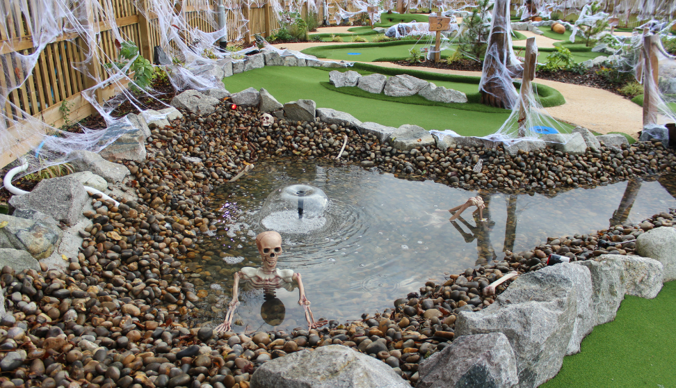 A Halloween-themed mini-golf course featuring artificial turf, rocks, and a pond with a fountain. Plastic skeleton decorations are positioned around and in the water, with one skeleton partially submerged as if emerging from the pond. White fake spider webs are draped along the wooden fence and posts, enhancing the spooky atmosphere. The mini-golf holes are surrounded by landscaping, rocks, and festive decor.